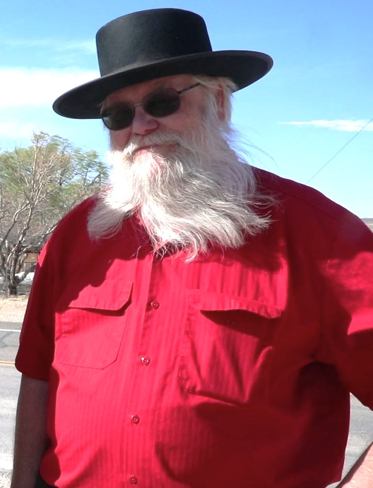 Historian Mark Hall-Patton in a desert setting wearing a black hat, sunglasses, and red shirt, with his famous white beard