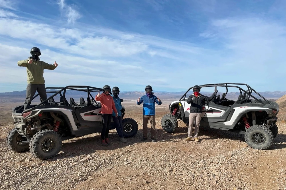 Helmeted riders give a thumbs-up beside two side-by-side UTVs on a desert ridge